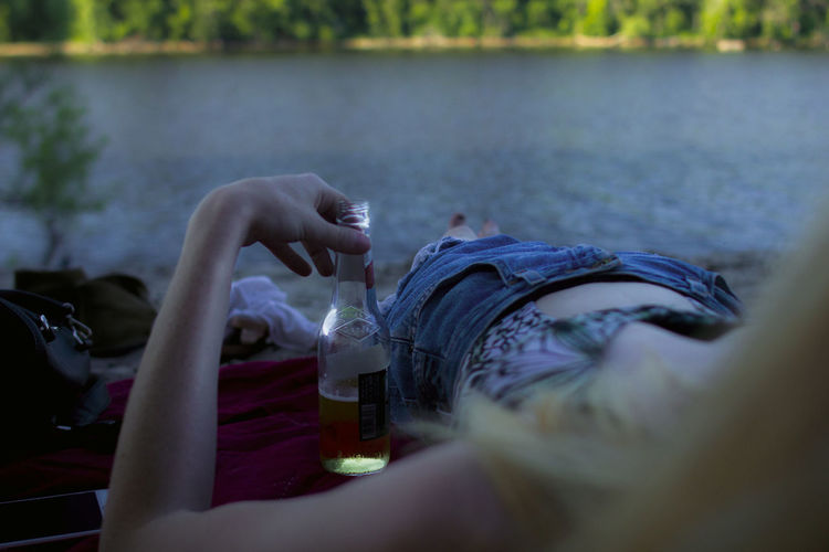 Low section of woman with beer bottle while relaxing at lakeshore