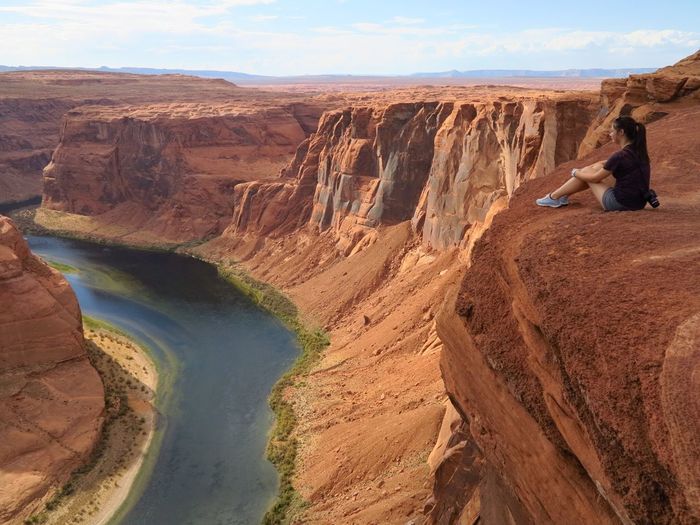 Full length of woman sitting on cliff by horseshoe bend against sky