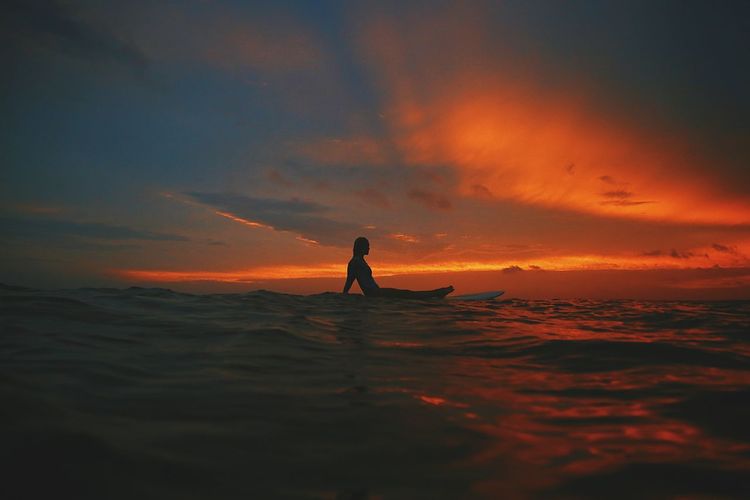 Silhouette young woman surfing on sea against cloudy sky during sunset