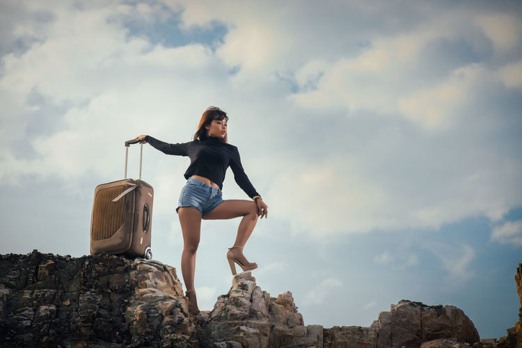 Low angle view of young woman with suitcase standing on rock formation against cloudy sky