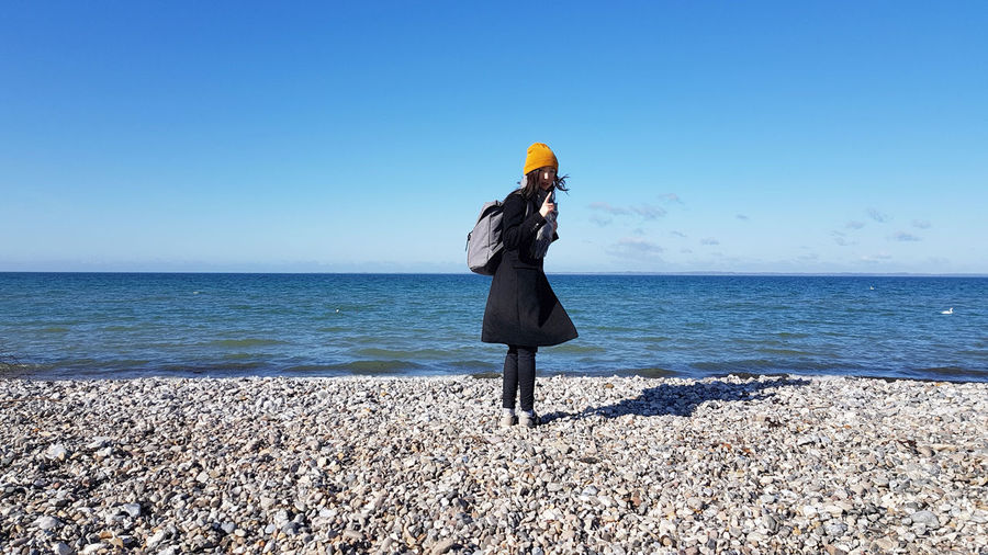 Portrait of backpack woman standing at beach against clear sky