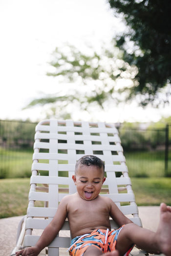 Cute shirtless baby boy sticking out tongue while sitting on chair against sky at water park
