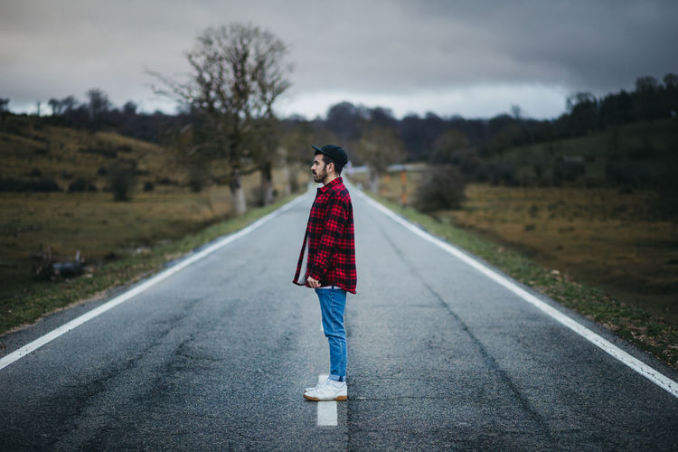Side view of man in casual wear walking on empty asphalt road among green fields with cloudy sky on background