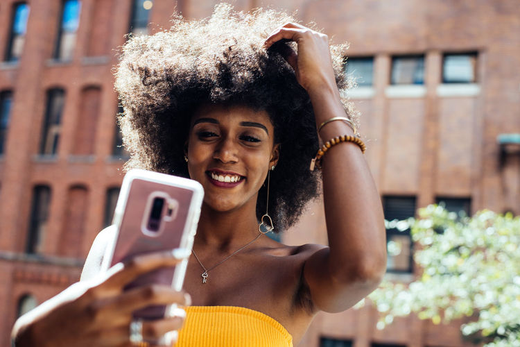 Happy young woman with afro hairstyle taking selfie through smart phone against building