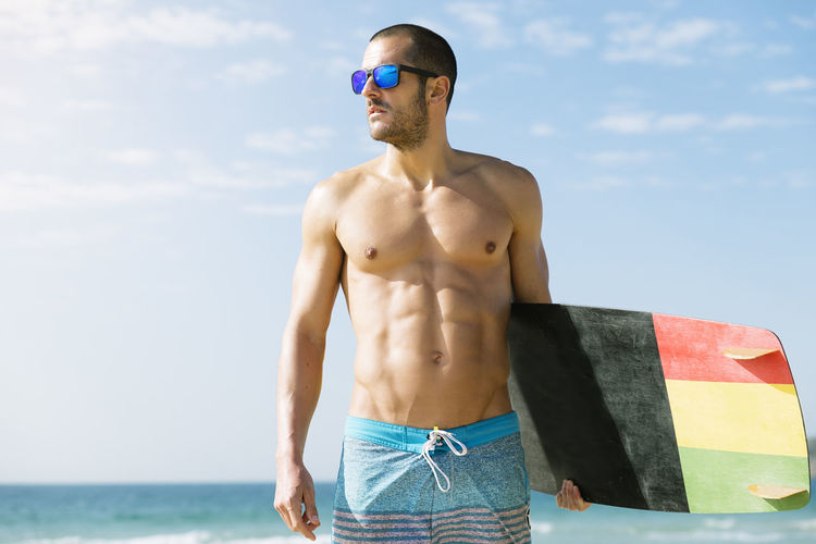 Shirtless man carrying surfboard against sky