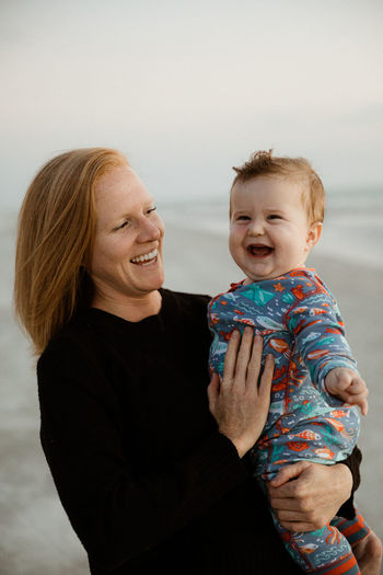 Single mom with red hair holds laughing chubby baby boy on the beach