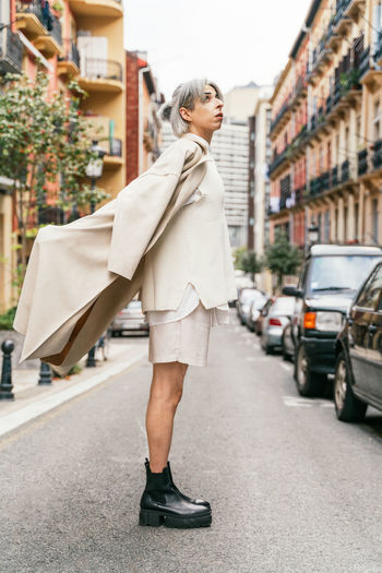 Side view of transgender female wearing trendy coat standing on road in city and looking up