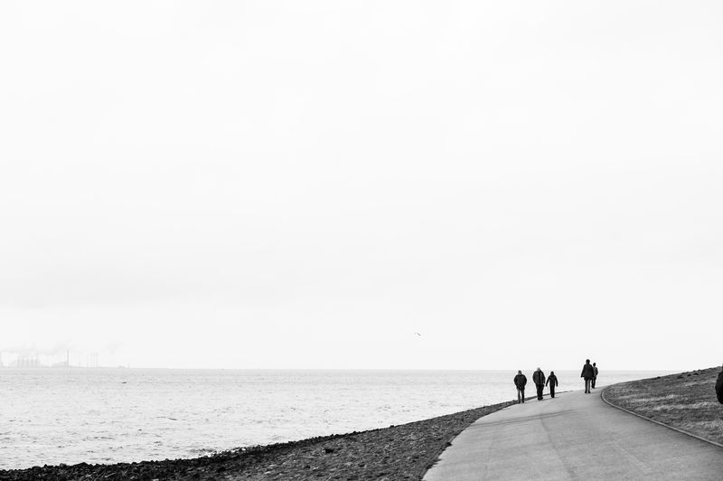 People walking on road by sea against clear sky