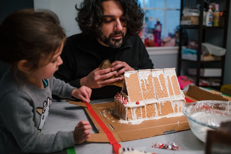 Father and daughter building gingerbread house at kitchen table