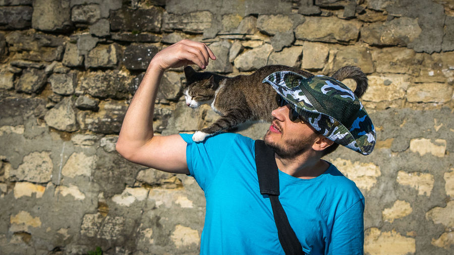 Man carrying cat on shoulder while standing by stone wall