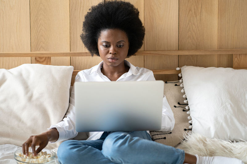 Woman using laptop while sitting on bed eating popcorn at home