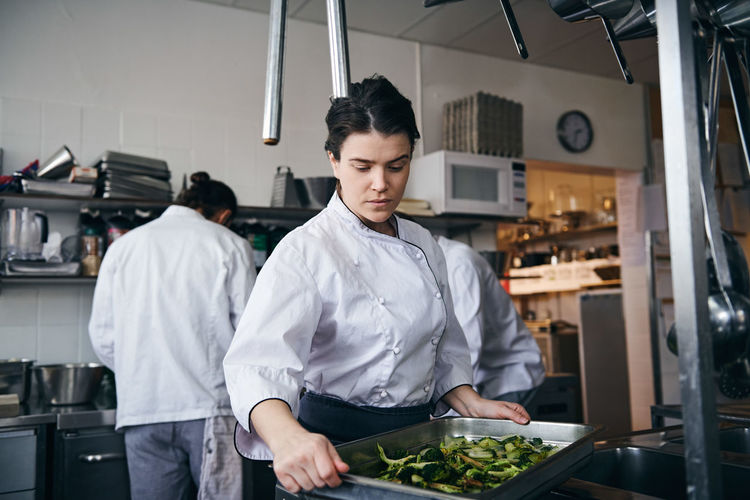 Mid adult female chef holding baking sheet with broccoli at commercial kitchen