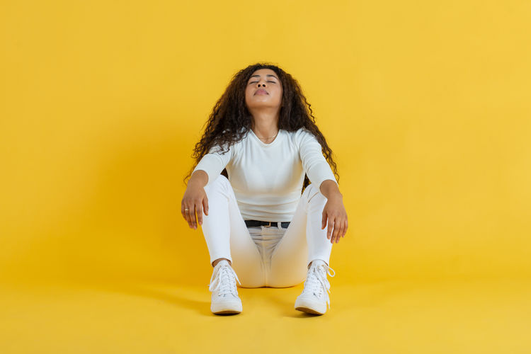 Portrait of young woman sitting on yellow background