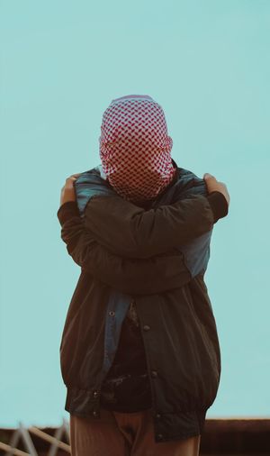 Man covering face with headscarf while standing against clear sky