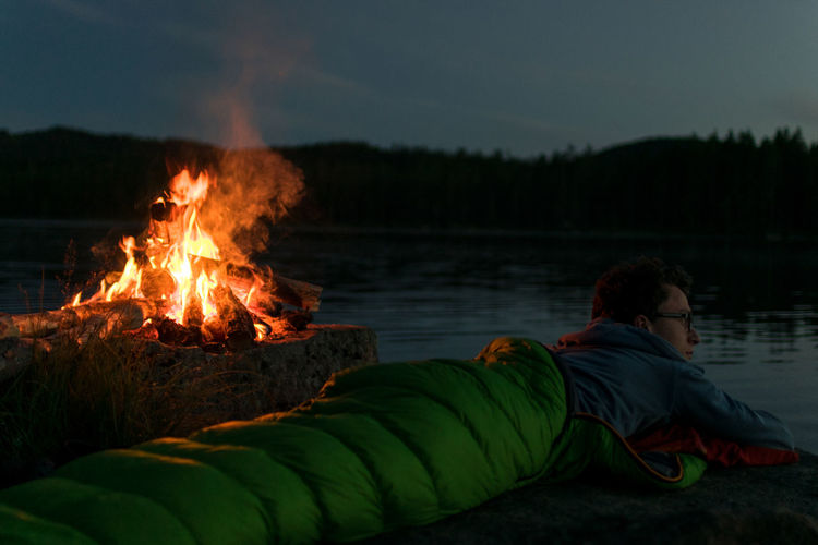 Man lying in sleeping bag by campfire at riverbank against sky