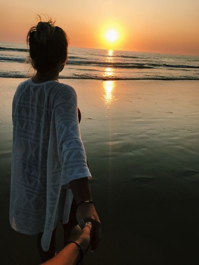 Woman holding cropped hand at beach against sky during sunset
