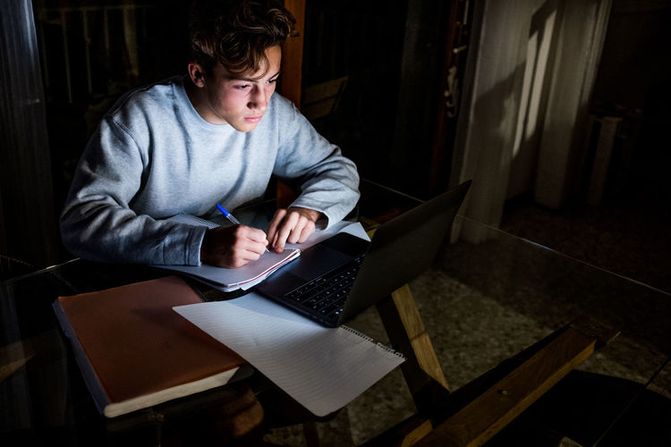 High angle view of teenage boy with laptop writing on book at home