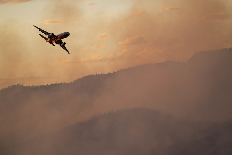 Military aircraft flying over smoke emitting from wildfire in forest during sunset