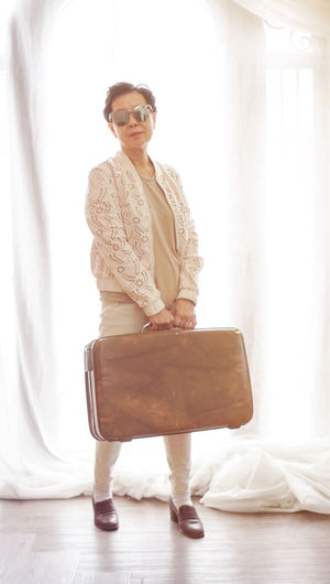 Portrait of woman holding suitcase while standing against window at home