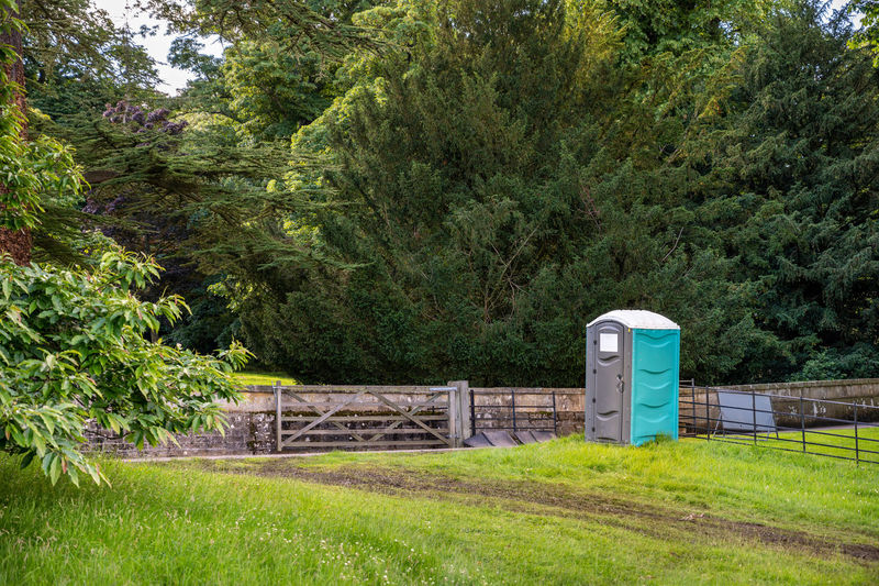 A plastic portable toilet next to muddy track in field at outdoor event and surrounded by tall trees