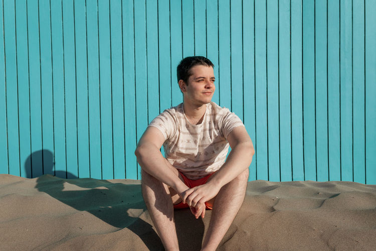 Young man with orange swimsuit pants looking at sun in sand beach
