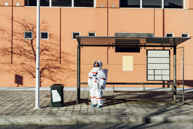 Young female astronaut in space suit waiting at bus stop during sunny day