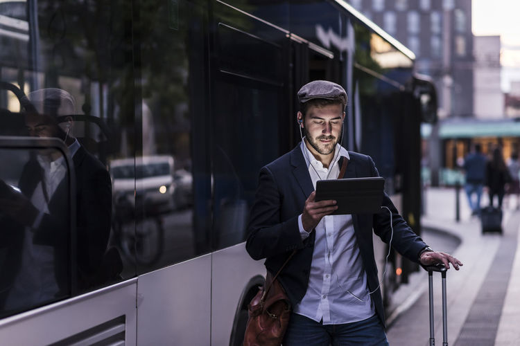 Young man at the bus stop in the city with earphones and tablet
