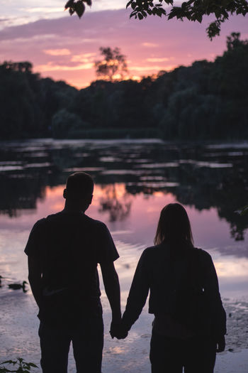 Rear view of couple standing by lake against sky during sunset