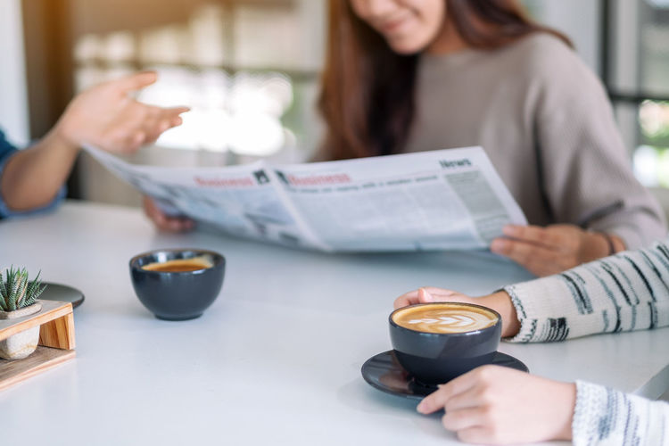 Midsection of woman holding newspaper by friends having coffee on laptop