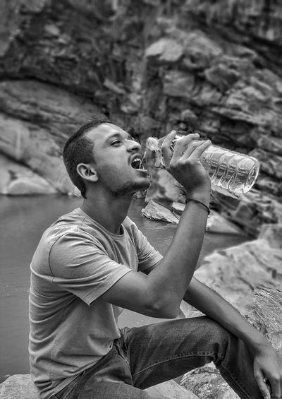 Man drinking water while sitting on rock at riverbank