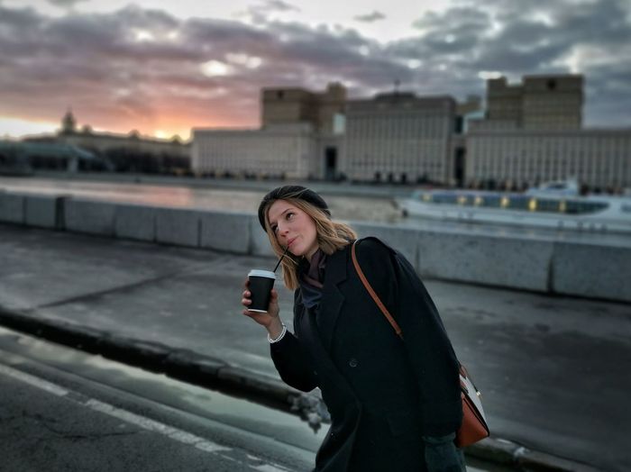 Woman drinking coffee while standing on road in city at sunset