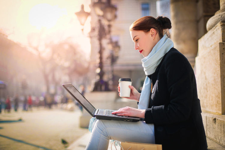 Woman using laptop while sitting on bench in city