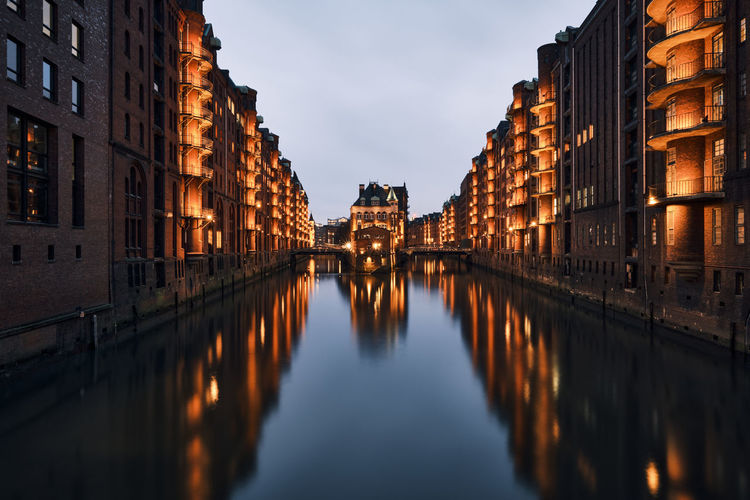Canal amidst illuminated city buildings against sky