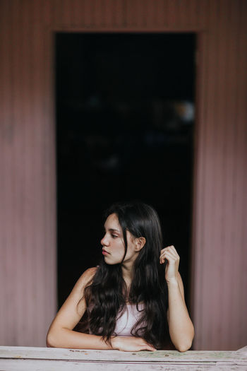 Calm dreamy female teenager with long dark hair standing looking away against plank wall