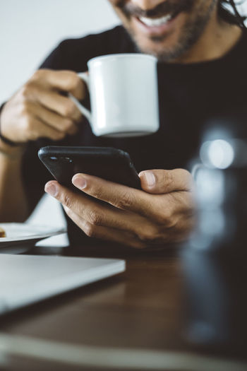 Midsection of man having coffee while using mobile phone at home