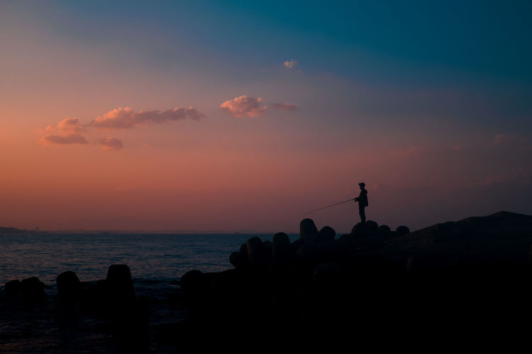 Silhouette people on rock by sea against sky during sunset