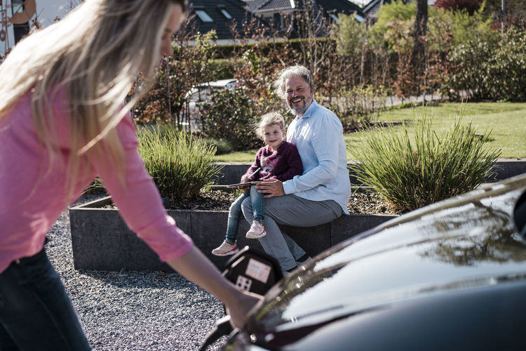 Smiling man with daughter looking at woman charging car on sunny day