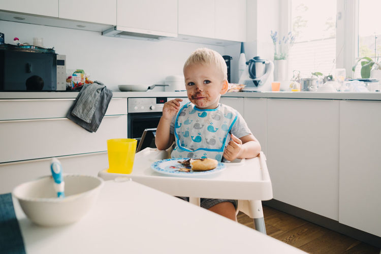 Adorable toddler with face covered in chocolate sitting on table at home