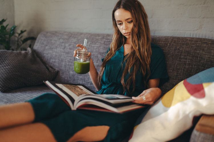 Young woman drinking juice while reading book on sofa at home
