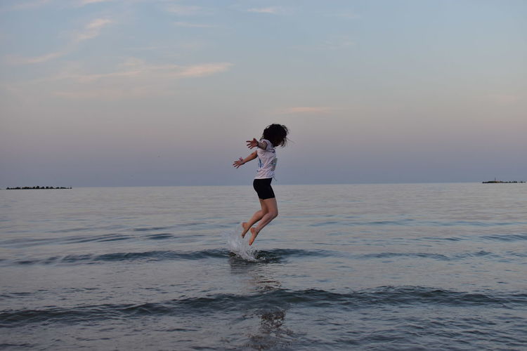 Side view of young woman jumping in sea against sky at sunset