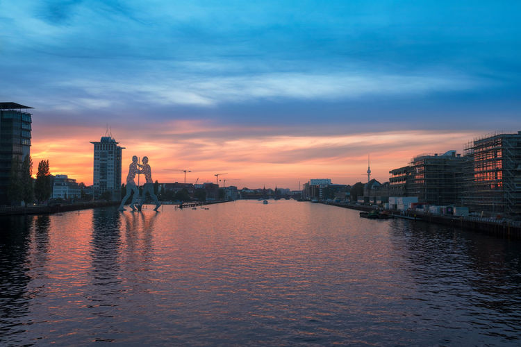 Scenic view of spree river by buildings against sky during sunset