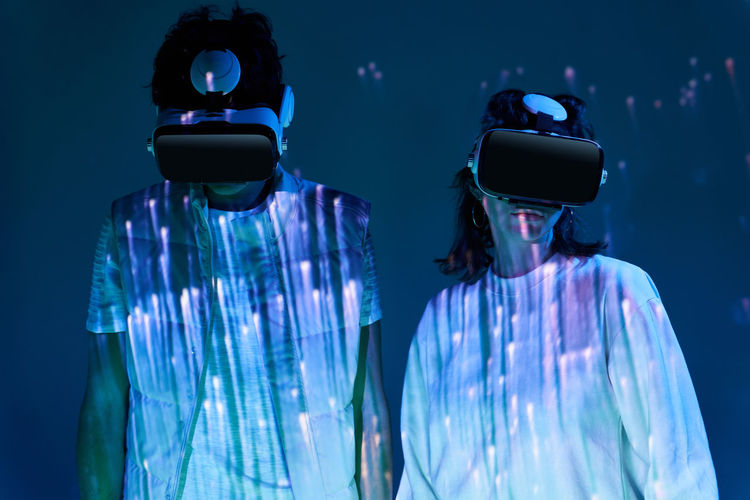 Couple in white wear and modern vr goggles standing in murk studio with glowing lights while exploring virtual reality together