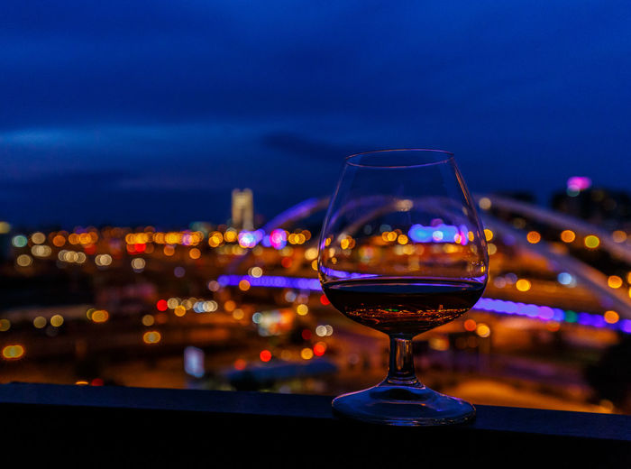 Close-up of wineglass on railing by illuminated cityscape against sky at night