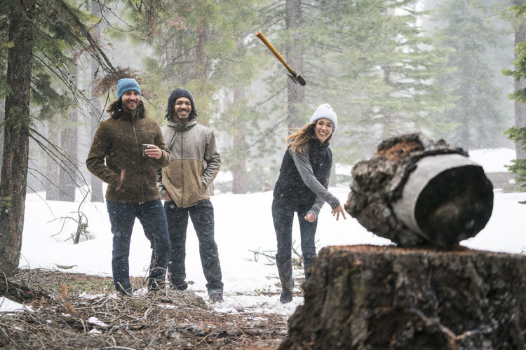 Young woman throws axe at a target in the snowy woods while her friends watch