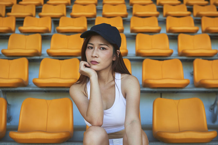 Confident asian athletic woman in sportswear sitting on stadium seat while resting after workout and looking at camera