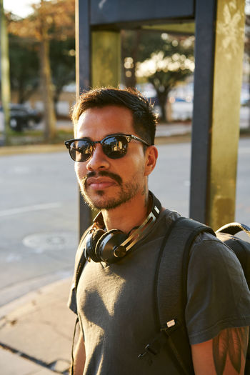 Young hispanic man in casual wear and stylish sunglasses with headphones and backpack looking at camera at city street