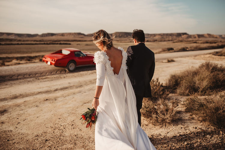 Back view of anonymous bride and groom strolling together towards luxury red car in desert of bardenas reales natural park in navarra, spain