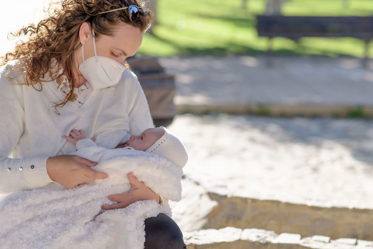 Mother wearing mask breastfeeding baby whiles sitting outdoors