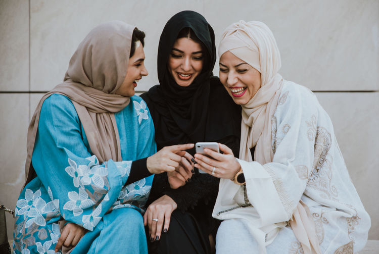 Cheerful friends in hijab looking at phone sitting against wall