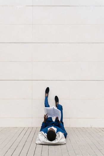 Young businessman lying with duffel bag on sidewalk while reading book in city